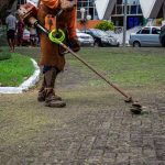 Trabalhador usa um aparador para fazer a manutenção da vegetação de um parque urbano, enquanto pedestres circulam pelo local.