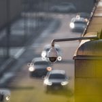 Câmera de monitoramento urbano voltada para uma via movimentada, com veículos desfocados ao fundo.