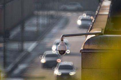 Câmera de monitoramento urbano voltada para uma via movimentada, com veículos desfocados ao fundo.