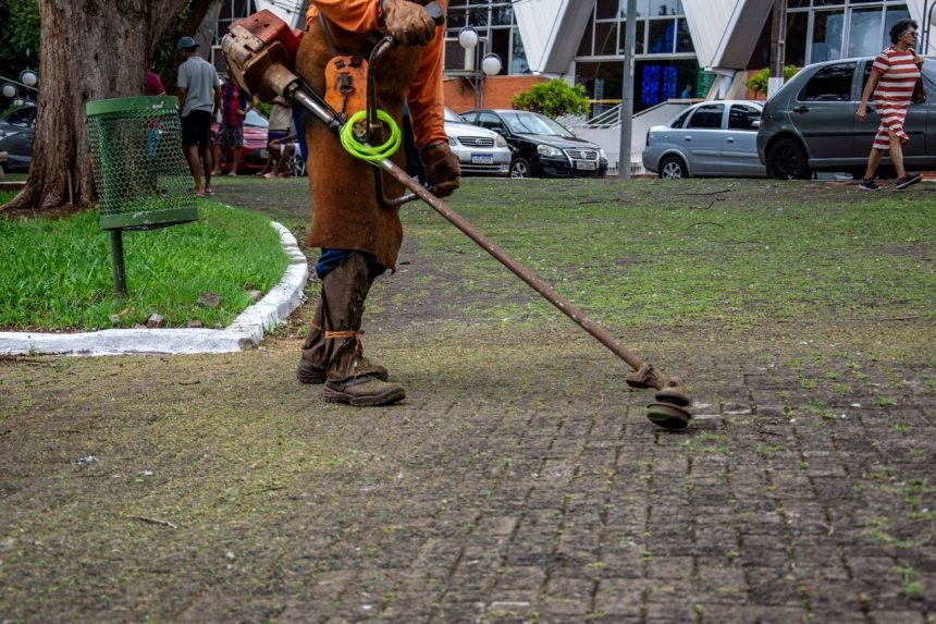 Trabalhador usa um aparador para fazer a manutenção da vegetação de um parque urbano, enquanto pedestres circulam pelo local.