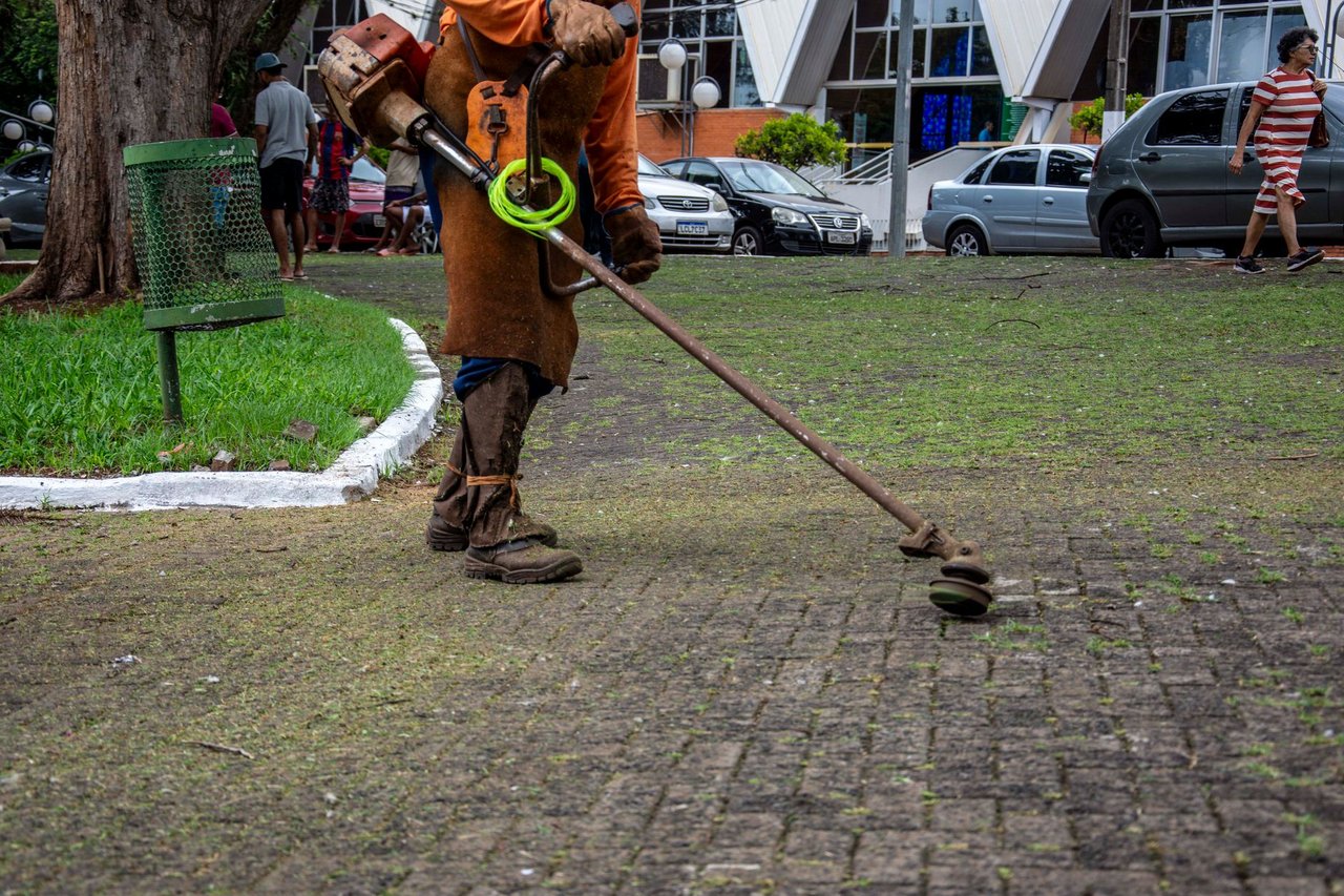 Trabalhador usa um aparador para fazer a manutenção da vegetação de um parque urbano, enquanto pedestres circulam pelo local.